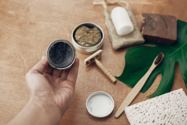 istock Zero waste. Hand holding natural eco charcoal toothpaste in glass jar on background of bamboo toothbrush, soap, solid shampoo, metal razor, deodorant on wood with green monstera leaf. 1126877116