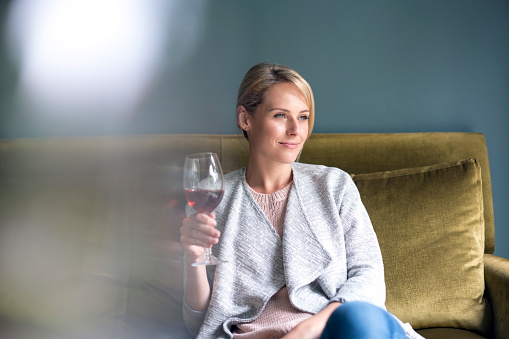 Smiling young woman holding red wine glass. Beautiful female is looking away. She is sitting on sofa at home.