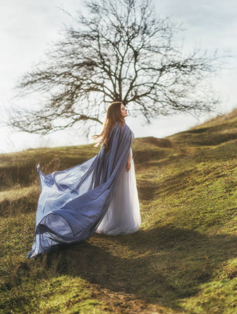 istock cute big plump model stands alone on a green summer hill in long gray and blue light, light dress and a flying flying cloak, mysterious attractive witch listening to the songs of the morning wind 1126891650