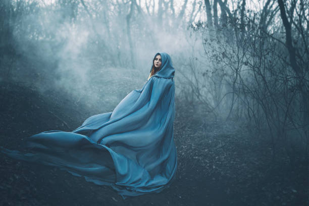 une mystérieuse sorcière noire se dresse seule dans une forêt dense et terrible, court intérieure de chasseurs, une peau mystérieux brouillard de fumée et blanc d’une femme dans un manteau de lumière vol long bleu. couleurs créatives - fairy forest women magic photos et images de collection