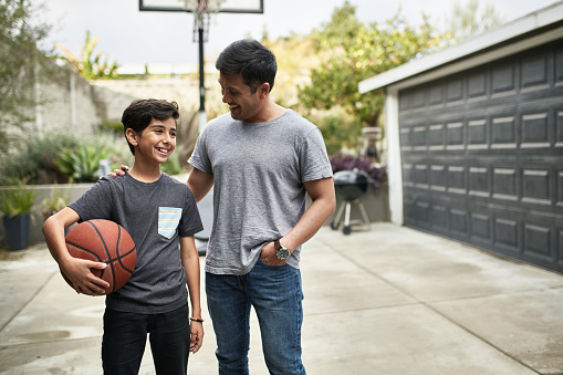 Father talking to smiling son with basketball. Happy mid adult man and child are standing in backyard. They are wearing casuals.