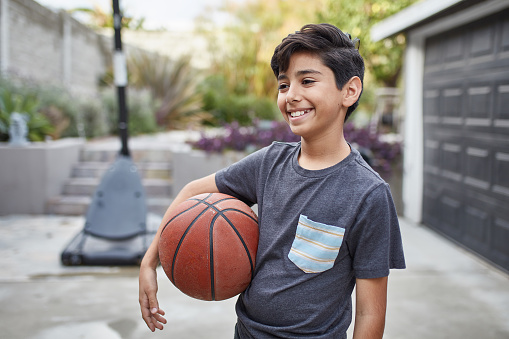 Boy looking away while holding basketball. Little male is smiling while enjoying weekend. He is wearing casuals in yard.
