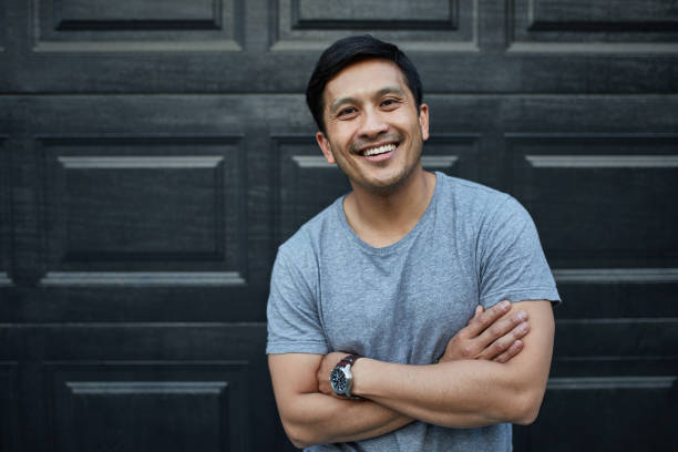 Happy man with arms crossed against wall at yard Portrait of smiling man with arms crossed. Happy mid adult male is standing against wall. He is in gray t-shirt at yard. hispanic guy stock pictures, royalty-free photos & images