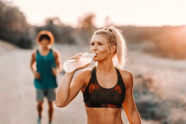 Photo of Beautiful blonde Caucasian woman drinking water while her boyfriend running behind her.