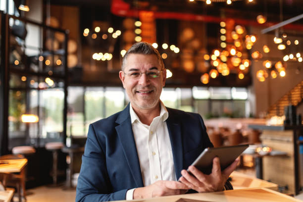 istock Middle aged man dressed smart casual holding tablet and looking at camera while sitting in cafeteria. 1126923245