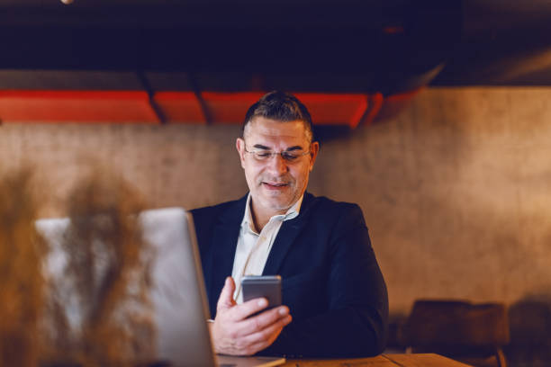 istock Middle aged businessman dressed smart casual using smart phone and sitting in cafeteria. In front of him on a desk laptop. 1126923481