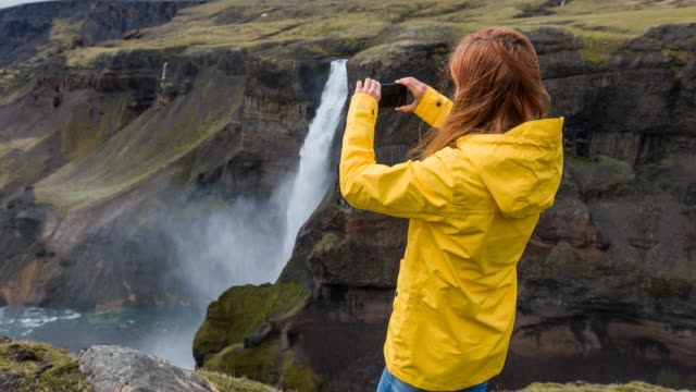 Woman standing on a cliff, taking pictures of Haifoss waterfall in Iceland with smartphone