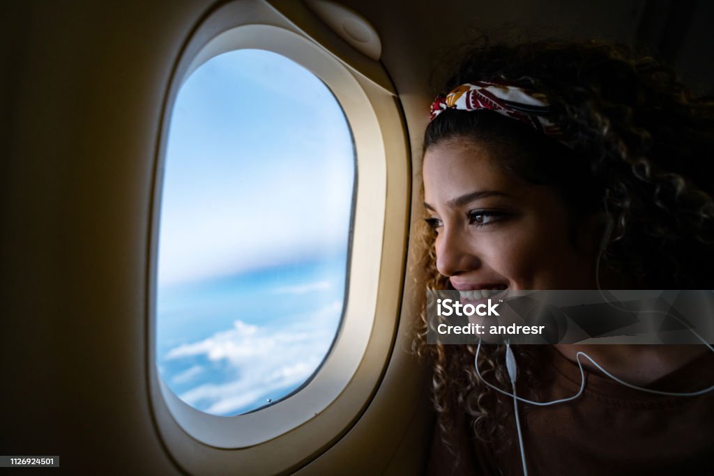 Woman traveling by airplane and listening to music Portrait of a happy woman traveling by airplane and listening to music with headphones while looking through the window - lifestyle concepts Airplane Stock Photo