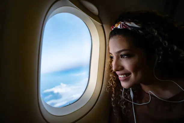 Portrait of a happy woman traveling by airplane and listening to music with headphones while looking through the window - lifestyle concepts