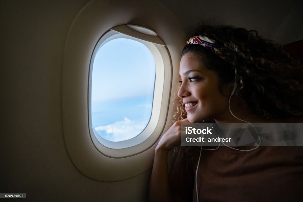 Woman traveling by plane and listening to music Portrait of a happy woman traveling by plane and listening to music with headphones while looking through the window - lifestyle concepts Airplane Stock Photo