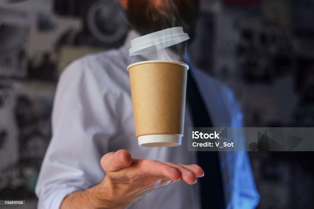 Barista and levitating paper cup of hot coffee Levitating in the air paper cup with hot coffee. Barista, a bearded young man in a white shirt with a tie, creates miracles - advertises his drink, causing it to soar. Logoplacement concept Adventure Stock Photo