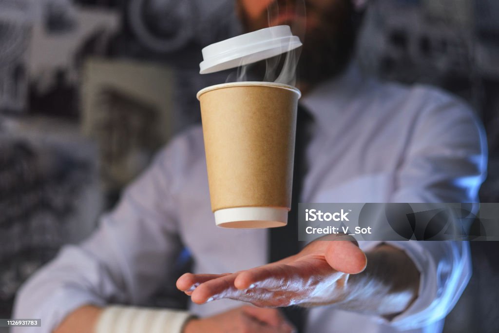 A waiter and a hovering cup of hot coffee Levitating in the air paper cup with hot coffee. Barista, a bearded young man in a white shirt with a tie, creates miracles - advertises his drink, causing it to soar. Logoplacement concept Adventure Stock Photo