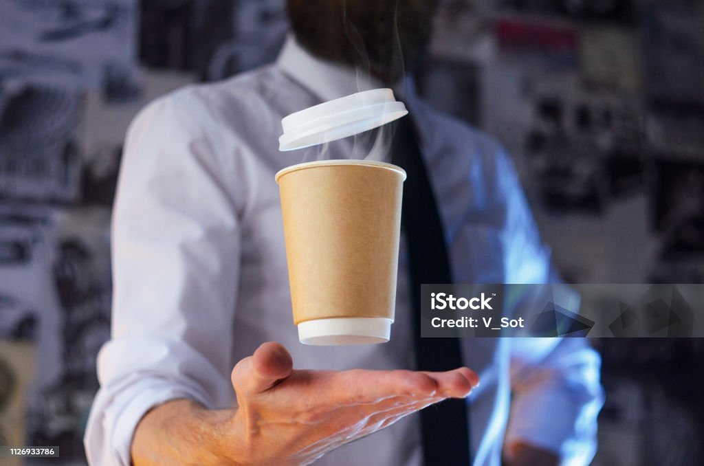 A waiter and a hovering cup of hot coffee Levitating in the air paper cup with hot coffee. Barista, a bearded young man in a white shirt with a tie, creates miracles - advertises his drink, causing it to soar. Logoplacement concept Adventure Stock Photo