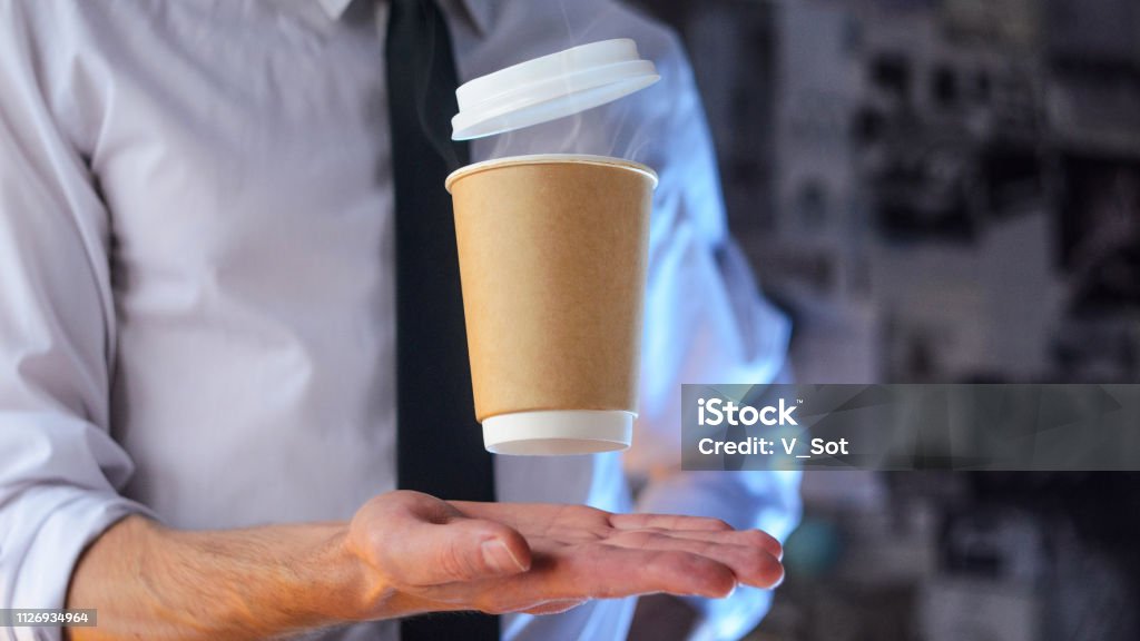 A waiter and a hovering cup of hot coffee Levitating in the air paper cup with hot coffee. Barista, a bearded young man in a white shirt with a tie, creates miracles - advertises his drink, causing it to soar. Logoplacement concept Adventure Stock Photo