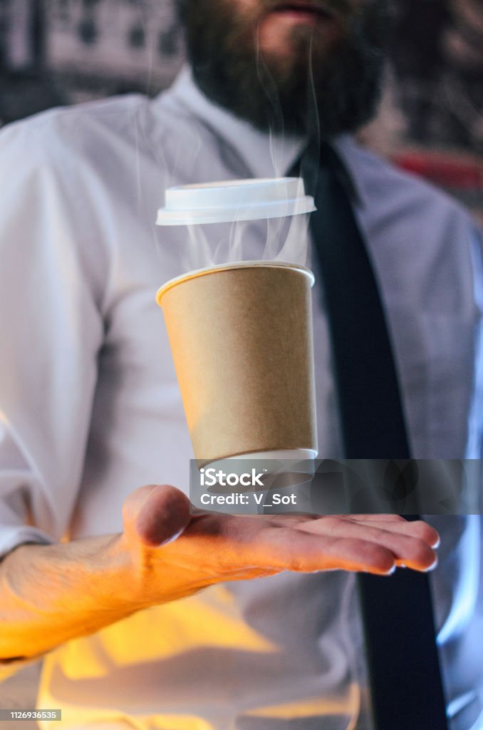 Barista and levitating cup of hot coffee Levitating in the air paper cup with hot coffee. Barista, a bearded young man in a white shirt with a tie, creates miracles - advertises his drink, causing it to soar. Logoplacement concept Adventure Stock Photo