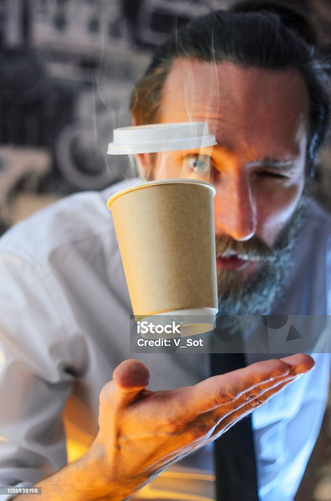 Levitate paper cup of coffee on the palm of a man Levitating in the air paper cup with hot coffee and evaporation from it. Barista, a bearded young man in a white shirt with a tie, creates miracles - advertises his drink, causing it to soar Illusion Stock Photo