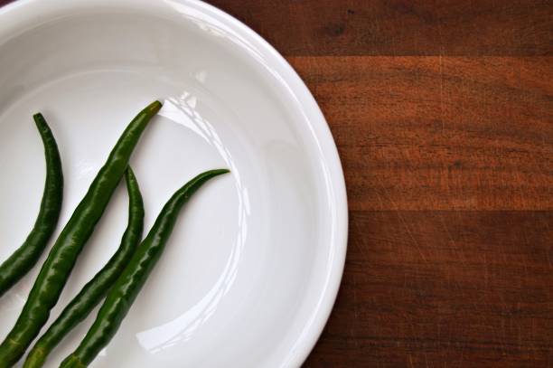 Horizontal photograph of close up of four green color chilli arranged in a circular white porcelain glass dish over a dark brown wooden board surface