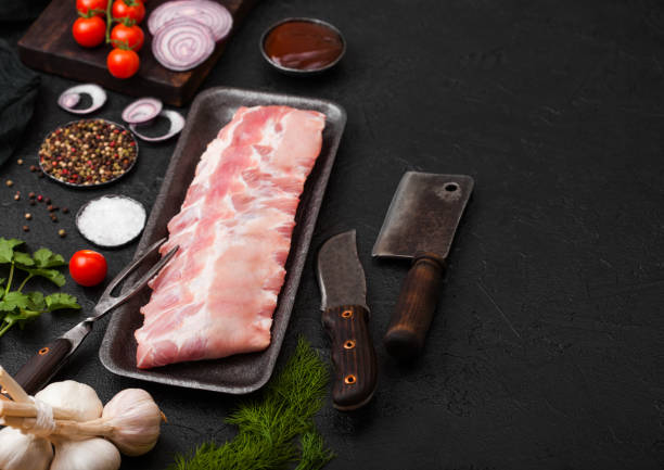 istock Plastic tray with raw pork ribs and vintage knife and fork on black stone background. Fresh tomatoes and red onion with garlic salt and pepper. Space for text 1126952348