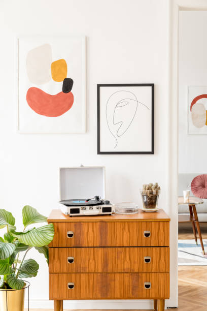 istock Vintage and retro living room with design commode, vinyl recorder and mock up posters frames on the white walls. Minimalistic room with brown wooden parquet and plants. 1126957932