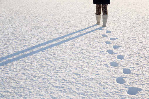 Young, adult woman's legs in white boots walking on fresh snow. Footprints behind human. Enjoying stroll in sunny, chilly winter day. Back view. Empty place for text, quote or sayings.