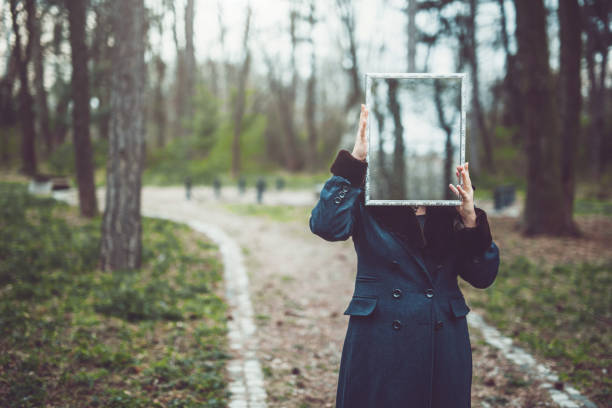 A woman in a green coat standing in a park area holding up a mirror that hides her face