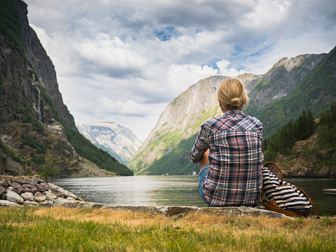 A woman sits on the shore of a picturesque fjord in Norway, admiring the beauty of Scandinavian nature.