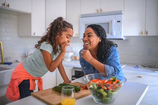 Smiling mother and daughter tasting vegetables at kitchen island. Woman and daughter preparing food together at home. They are representing healthy lifestyle.