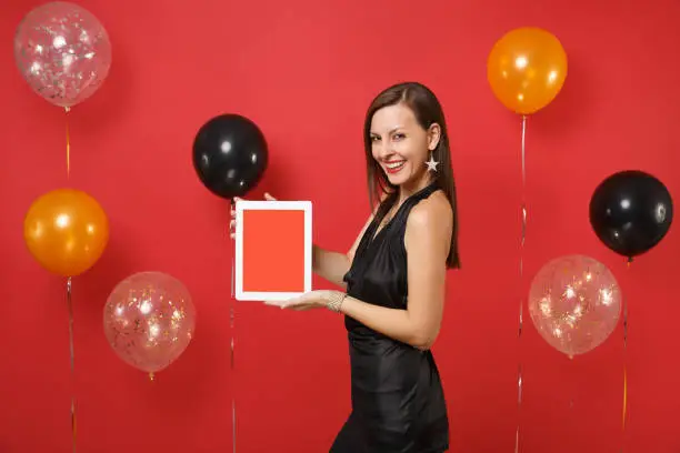 Photo of Seductive young girl in black dress celebrating, holding tablet pc computer with blank black empty screen on bright red background air balloons. Happy New Year, birthday mockup holiday party concept.