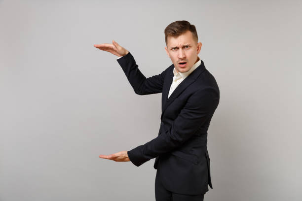 istock Concerned young business man in classic black suit gesturing demonstrating size with vertical copy space isolated on grey wall background. Achievement career wealth business concept. Advertising area. 1126986477
