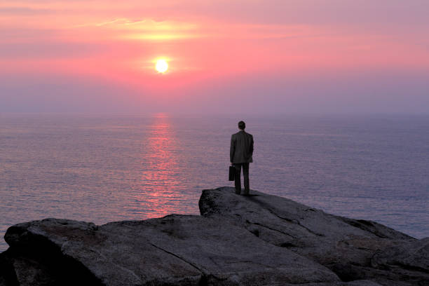 A businessman stands with his back to the camera while holding his briefcase as he stands on top of a rocky cliff and looks out as the sun is about to set into the ocean.