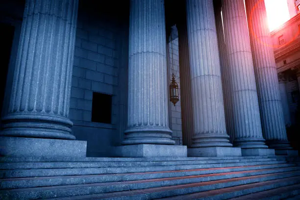 The sun peaks out from above a courthouse as red and blue tones envelope the Greek columns of the portico.
