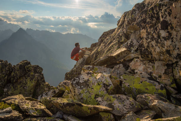 A young man rock climbing on the top of a mountain above a steep, forested valley.