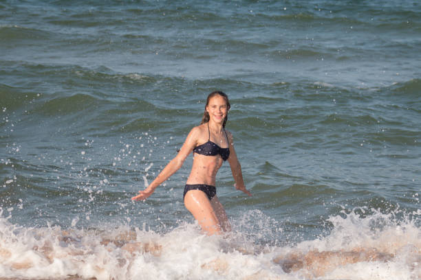 летом красивая молодая девушка-подросток с длинными волосами, 13-14 лет, резвится на пляже в пене волн у берега моря - 13 14 years стоковые фото и изображения