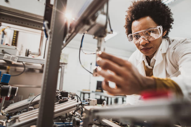 Black female engineer working on industrial machine in a laboratory. Low angle view of African American lab worker examining machine part while working in a lab. quality control photos stock pictures, royalty-free photos & images