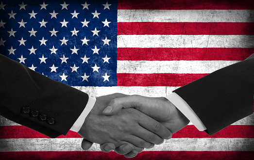 Two men/politicians in suits shaking hands with the national flags of the United States of America on the background