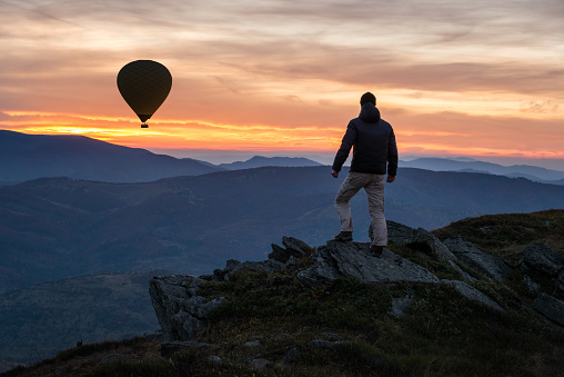 Traveler on mountain peak at sunset, hot iar balloon flying away