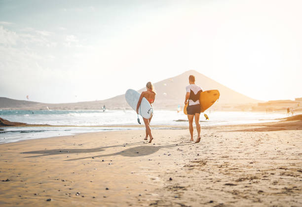 glückliche surfer mit surfbrettern auf dem strand - sportliches paar spaß surfen zusammen bei sonnenuntergang - extreme beziehung, menschen, sport und jugend lifestyle-konzept - bonding horizontal surfing surf stock-fotos und bilder