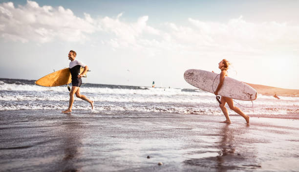 glückliche surfer mit surfbrettern am meer entlang laufen ufer - sportliche paare, die spaß gehen zusammen bei sonnenuntergang - extreme surfen sport, sportliche leute und jugend-beziehung-lifestyle-konzept zu surfen - bonding horizontal surfing surf stock-fotos und bilder