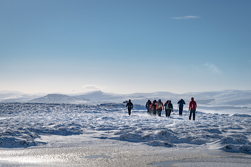 Group of hikers walking in the snow across a vast beautiful wild scenic landscape, with rucksacks and poles, with sunny sky on a winter day, with frozen pool of water in foreground and distant mountain ridges, shot in County Wicklow, Ireland