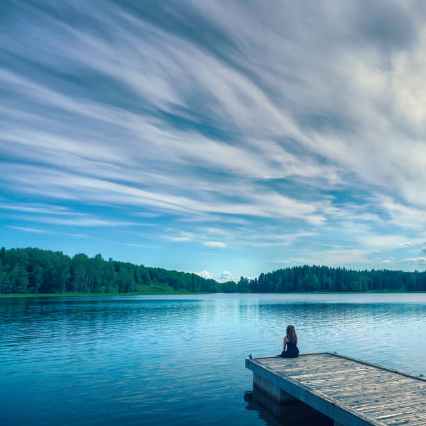 Alone by the lake A woman sits on the edge of a jetty at peaceful lake with a dramatic sky. swedish woman stock pictures, royalty-free photos & images