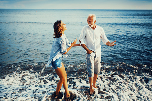 Grandfather telling jokes to his granddaughter while they walking on the beach during family summer vacation. They are laughing and enjoying together.