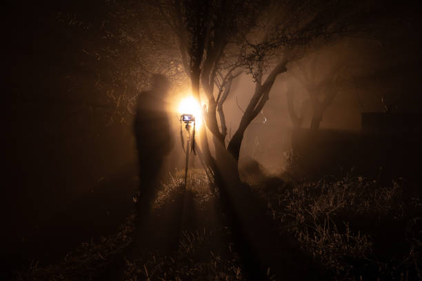 istock Foggy night yard with bright light on background. lantern hanging on the tree and silhouette of cameraman with his camera. 1127138446
