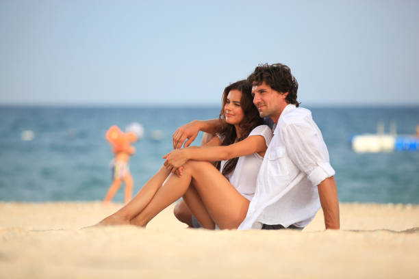 istock Happy young couple sitting on beach and pleasuring and smiling while sunbathing 1127153156