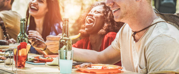 Happy multiracial friends eating, drinking beer and laughing together at barbecue dinner - Young happy people having fun at bbq meal - Friendship and food concept - Focus on right man mouth Happy multiracial friends eating, drinking beer and laughing together at barbecue dinner - Young happy people having fun at bbq meal - Friendship and food concept - Focus on right man mouth south african braai stock pictures, royalty-free photos & images