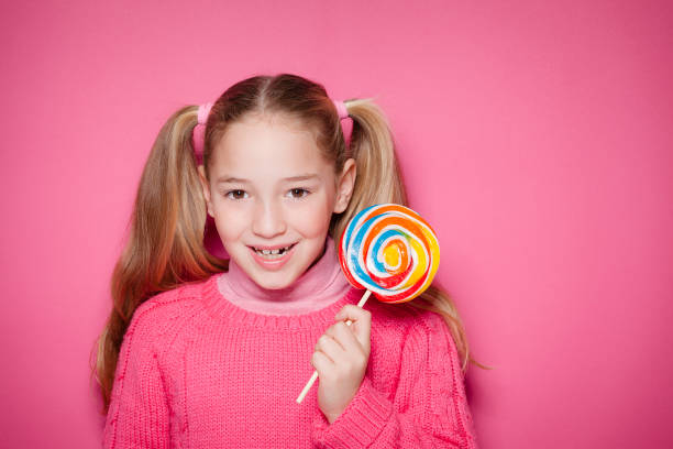 A young Asian woman with short brown hair is smiling while holding a large red and white spiral lollipop. She is wearing a white, short-sleeved top and a gray skirt, and a light brown straw hat. The image is a studio portrait against a plain white background, highlighting the woman's cheerful expression and the vibrant color of the lollipop.