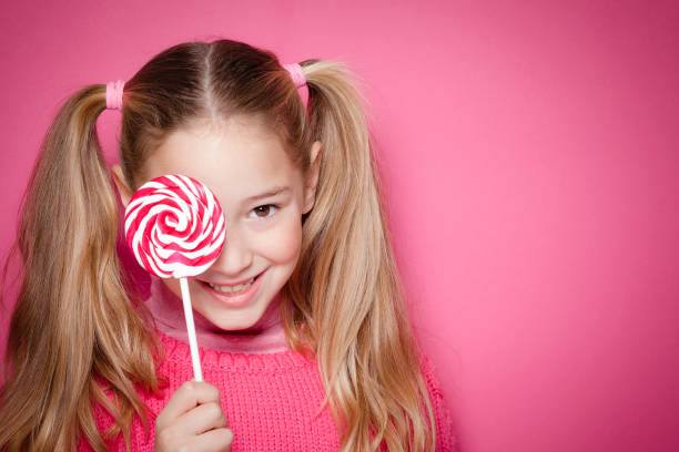 Blonde young girl with pigtails holding red and white spiral lollipop in front of the right eye and looking at camera with the left one shot against pink background. The girl is wearing a pink sweater. The composition is at the left leaving useful copy space for text and/or logo at the right. Predominant color is pink. DSRL studio photo taken with Canon EOS 5D Mk II and Canon EF 70-200mm f/2.8L IS II USM Telephoto Zoom Lens