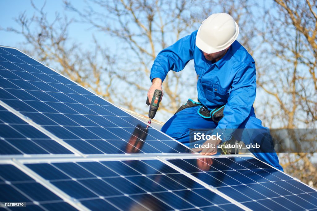 Electrician mounting solar panel on roof of modern house Male engineer in blue suit and protective helmet installing photovoltaic panel system using screwdriver. Professional electrician mounting solar module on roof. Alternative energy ecological concept. Solar Panel Stock Photo