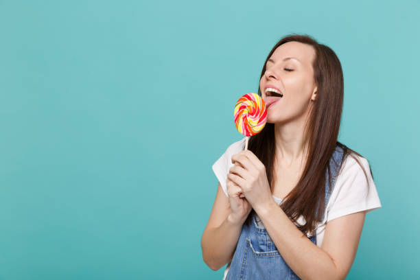 Portrait of cute young woman in denim clothes with closed eyes hold, licking colorful round lollipop isolated on blue turquoise wall background in studio. People lifestyle concept. Mock up copy space retrato de mujer joven linda en ropa del dril de algodón con ojos cerrados hold, lamiendo colorido chupetín redondo aislado sobre fondo de pared azul turquesa en estudio. concepto de estilo de vida de las personas. se burlan de espacio de copia.