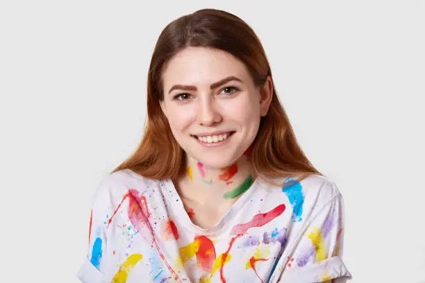 Close up shot of happy smiling talented painter has dark straight hair, traces of paints on clothes, recieves good remarks of her work, looks directly at camera, isolated on white background