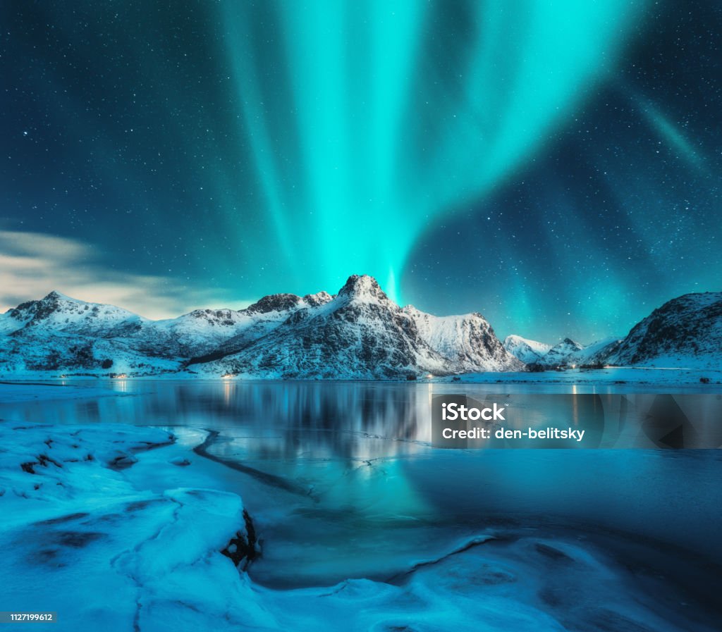 Aurora borealis over snowy mountains, frozen sea coast, reflection in water at night. Lofoten islands, Norway. Northern lights. Winter landscape with polar lights, ice in water. Starry sky with aurora Landscape - Scenery Stock Photo